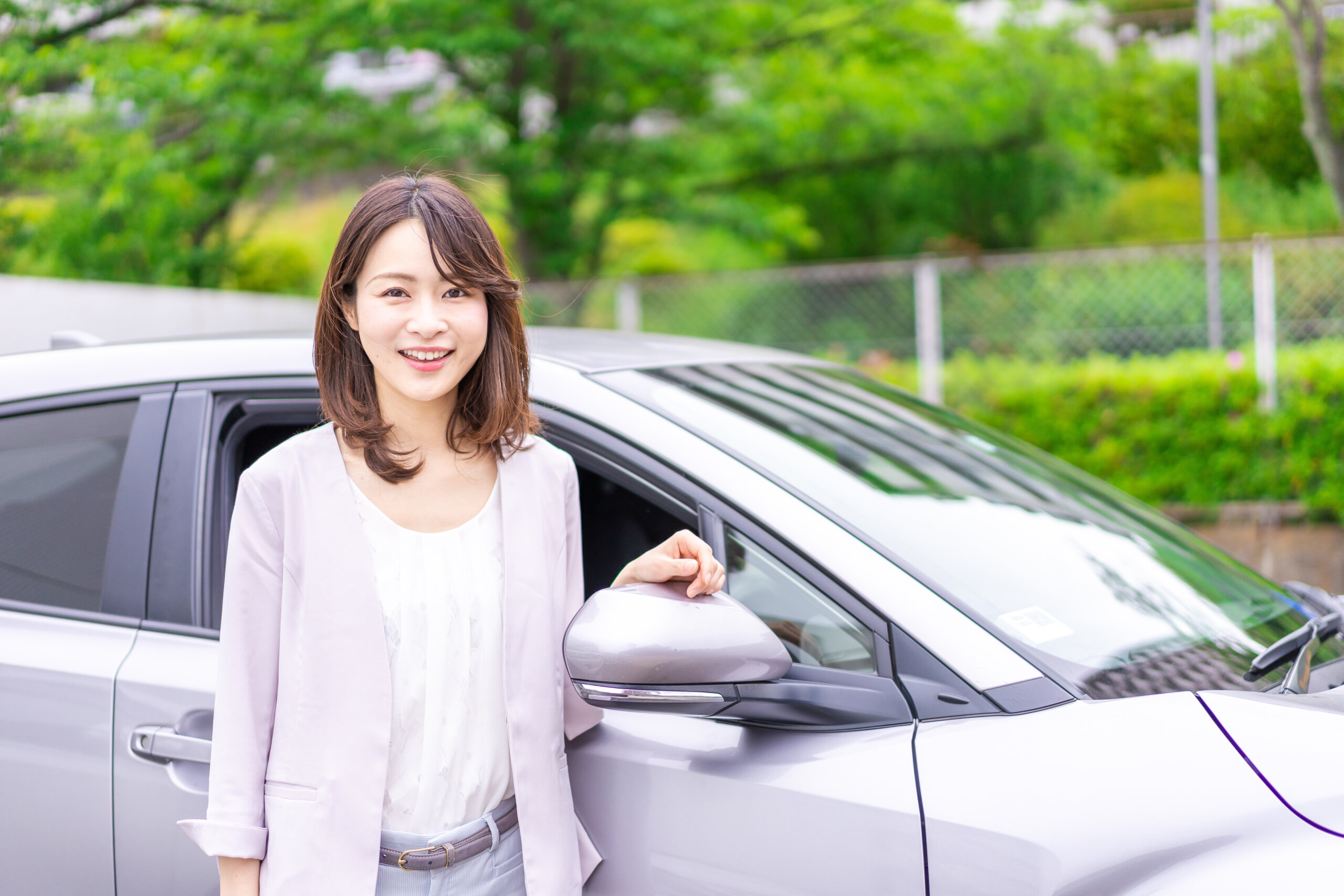 Smiling woman stands beside a silver car with the door open, in a green outdoor area.