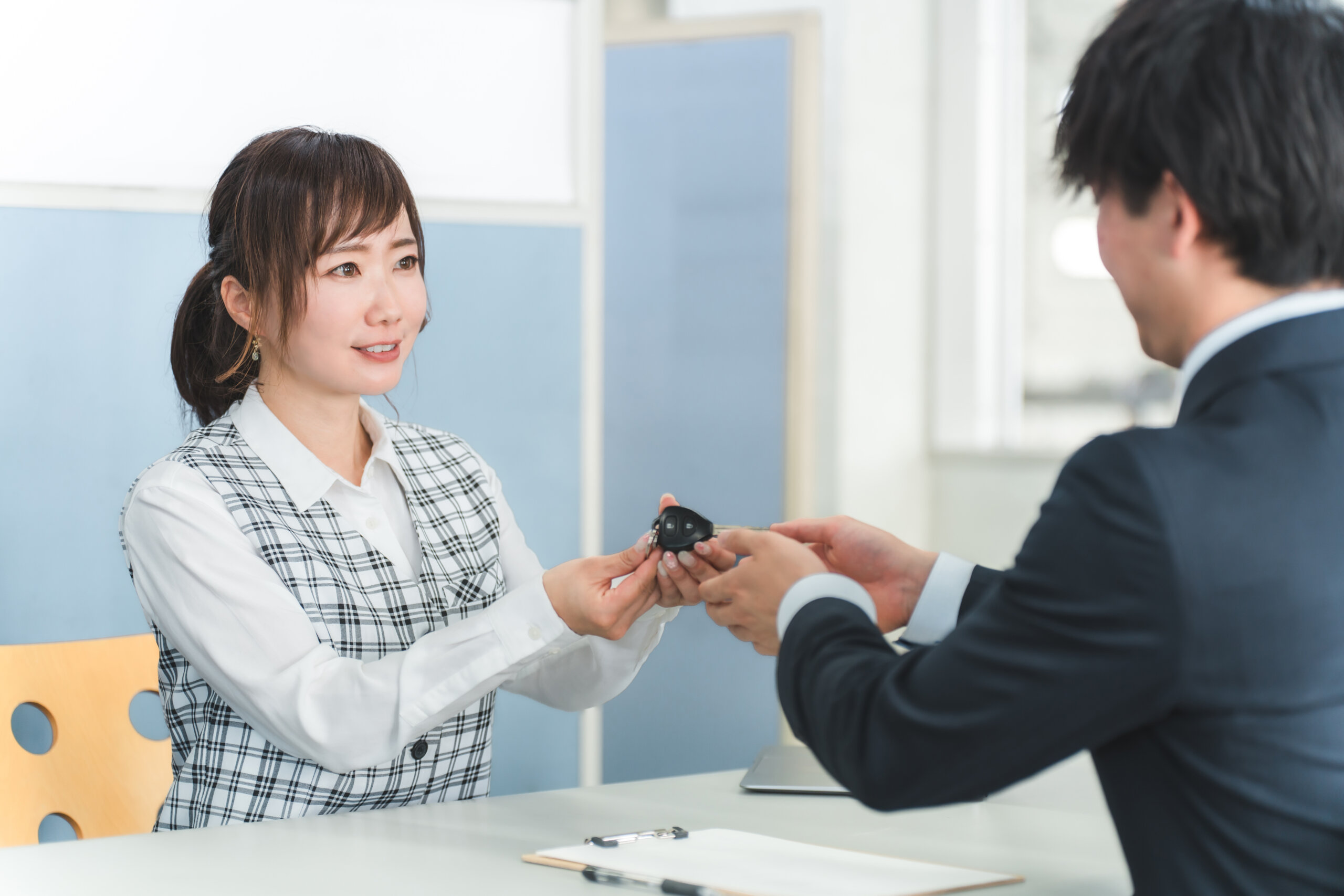 Woman in plaid blazer hands over car key fob to a man across a desk, in a bright office setting.