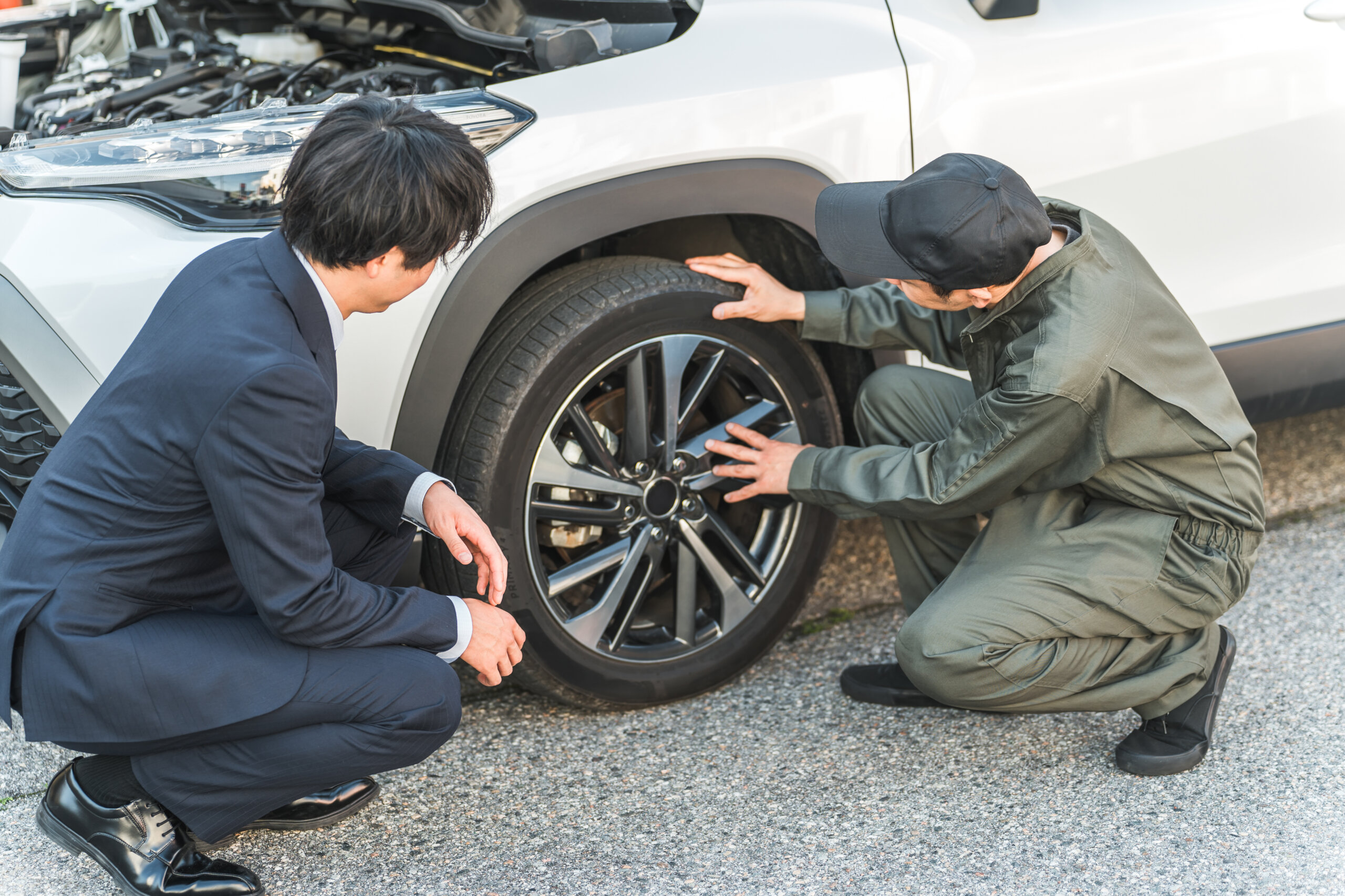 Two men crouch beside a white SUV, inspecting a front tire with its hood open in the background.