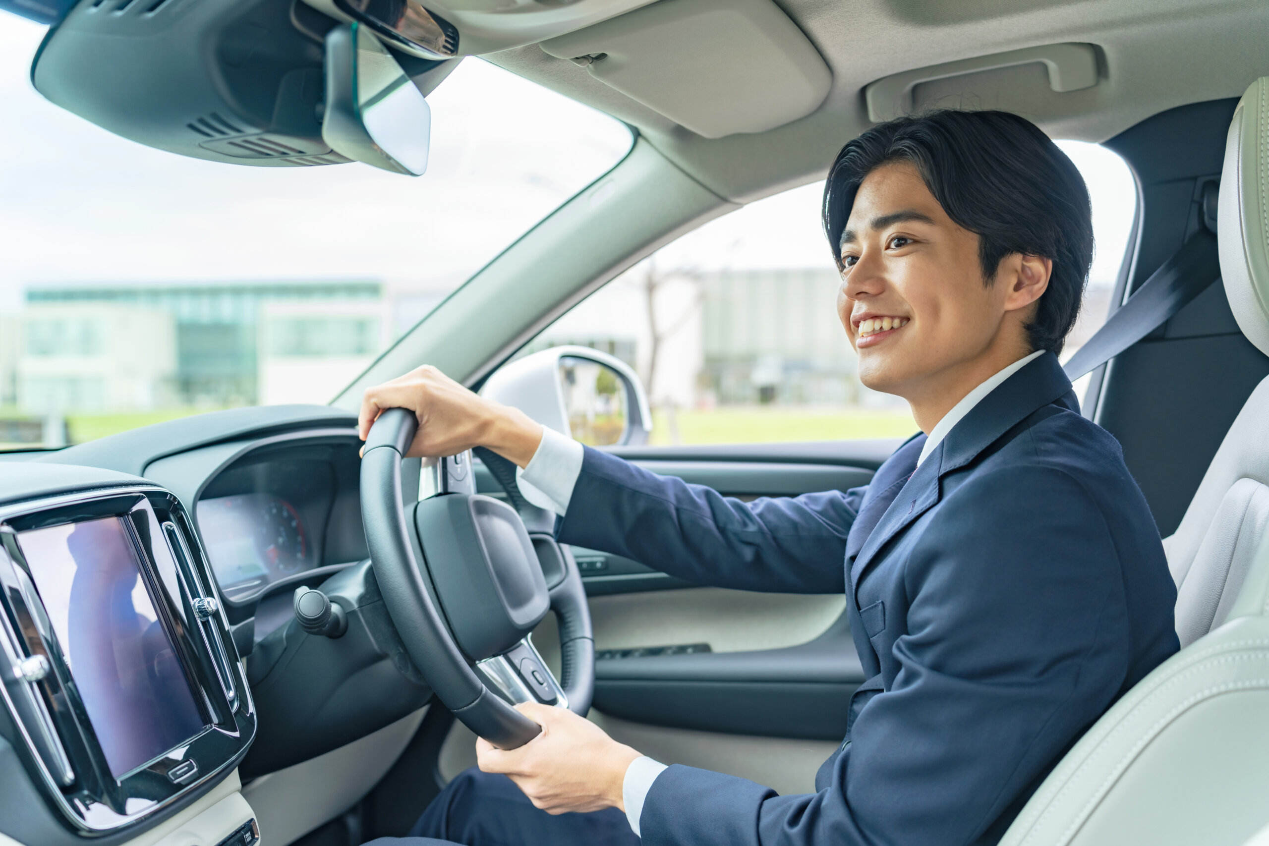 Smiling man in a suit drives a car, hands on the steering wheel inside a modern vehicle.